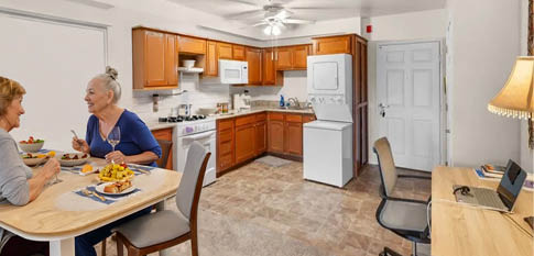 Two women are sitting at a table in a kitchen, eating food. The kitchen is equipped with a refrigerator, oven, sink, and dining table. There are chairs around the table, and a laptop is placed on the table. AI generated content